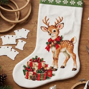 Könnte beinhalten: Ein weißer Weihnachtsstrumpf mit einem Kreuzstichmotiv eines braunen Rehkitzes, das eine rote Schleife mit Glöckchen trägt und neben einem Stapel Geschenke steht. Der Strumpf hat ein grünes Bündchen mit weißen Schneeflockenmustern.