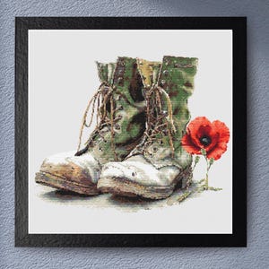 May include: A framed print featuring a pair of worn military boots with camouflage detailing, brown laces, and a red poppy. The boots are off-white and green, with visible wear and tear. The poppy is in full bloom.