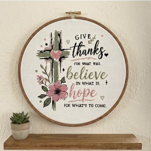 May include: A circular wooden embroidery hoop containing white fabric with a cross-stitch design. The design features a green cross, a pink heart, and pink flowers. It includes the text: GIVE thanks FOR WHAT WAS. believe IN WHAT IS. hope FOR WHAT'S TO COME.