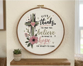 May include: A circular wooden embroidery hoop containing white fabric with a cross-stitch design. The design features a green cross, a pink heart, and pink flowers. It includes the text: GIVE thanks FOR WHAT WAS. believe IN WHAT IS. hope FOR WHAT'S TO COME.