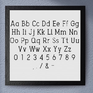 Op de afbeelding: Een ingelijste print met het alfabet en cijfers in een schreeflettertype. De zwarte lijst omsluit een witte achtergrond met zwarte letters en cijfers. De letters zijn in rijen gerangschikt, met hoofdletters en kleine letters, en de cijfers 0-9.