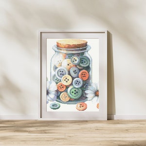 Puede incluir: Un frasco de vidrio lleno de botones coloridos, incluyendo azules, verdes, blancos y naranjas. El frasco está sentado sobre una superficie blanca con flores blancas.