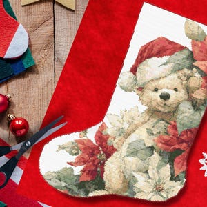 Puede incluir: Un calcetín navideño con un oso de peluche con un gorro de Papá Noel, rodeado de flores de Pascua. El calcetín está sobre un fondo rojo con adornos navideños, fieltro y tijeras.