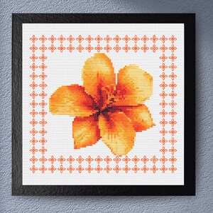 May include: Cross-stitch art of a vibrant orange flower with detailed petals, framed in a black square. The flower is centered on a white background, surrounded by an orange floral border. The artwork is displayed in a black frame.
