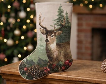 Op de afbeelding: Een kerstkous van geweven stof met een gedetailleerde afbeelding van een hert met gewei in een bos van groene dennenbomen. De onderkant van de kous is versierd met een patroon van dennenappels en rode hulstbessen.