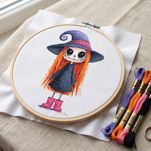May include: A cross-stitch embroidery hoop featuring a cartoon witch with orange hair, a purple hat, and a black dress. The embroidery is on white fabric, held in a wooden hoop. Colorful embroidery floss and small scissors are next to the hoop.