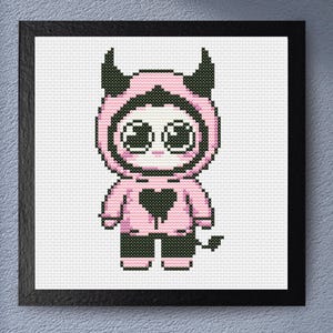 May include: A framed cross-stitch artwork featuring a cute devil character. The character is depicted in a pink hooded outfit with black horns, a heart, and a tail. The artwork is framed in a black square frame.