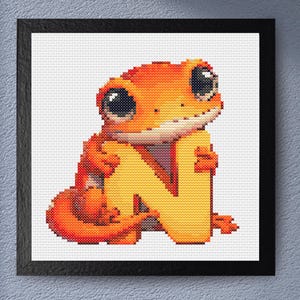 May include: Cross-stitch art of an orange gecko holding a yellow letter "N". The gecko has large black eyes and is set against a white background. The artwork is framed in a black square frame, suitable for nursery decor.
