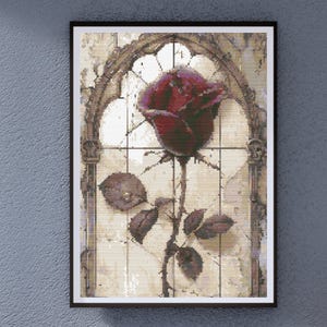 May include: A framed cross-stitch art piece featuring a large, dark red rose in full bloom. The rose is set against a backdrop of a stained-glass window design with a gothic arch. The artwork has a vintage, antique aesthetic.
