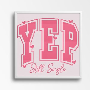 May include: A framed cross-stitch artwork with the word "YEP" in large pink letters, accented with small hearts. Below, the phrase "Still Single" is stitched in a cursive font. The artwork is set against a light pink background.