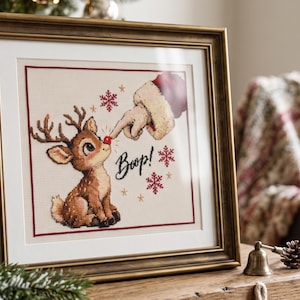 May include: A framed cross-stitch artwork featuring a brown reindeer with a red nose being touched by a gloved hand. The text "Boop!" is written in black script, surrounded by red and gold snowflake icons on a cream-colored fabric background.