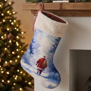 Peut inclure: Une chaussette de Noël ornée d'une scène hivernale avec le Père Noël marchant dans une forêt enneigée. La chaussette a un bord en fausse fourrure blanche et une boucle de suspension rayée rouge et blanche. Un sapin de Noël illuminé est en arrière-plan.