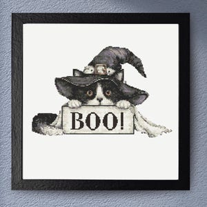 May include: A framed cross-stitch embroidery featuring a black and white cat wearing a dark pointed witch hat. The cat is peeking over a rectangular sign that has the word BOO! printed in black capital letters on a light background.