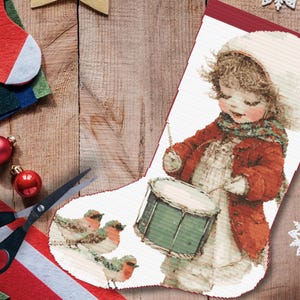 May include: A Christmas stocking featuring a vintage illustration of a child playing a drum, wearing a red coat and white hat. The stocking is decorated with a red border and a design of birds. Additional Christmas decorations are visible.