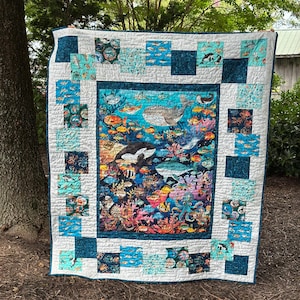 May include: A colorful quilt featuring an underwater scene with whales, orcas, and various fish. The quilt has a white border with teal and patterned squares. The central image showcases vibrant coral and marine life.