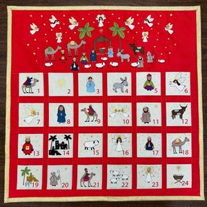 May include: Red Advent calendar with embroidered nativity scene and numbered squares. The calendar features angels, camels, and other figures. Each square is numbered from 1 to 24, with a gold border.