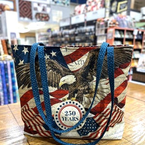 May include: A quilted tote bag featuring a patriotic design with a bald eagle, American flag motifs, and a circular emblem that reads 250 YEARS. The bag has two blue fabric straps patterned with small white stars and a diamond-stitched texture.