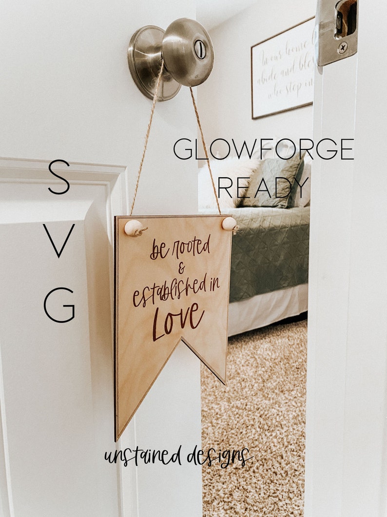 May include: A wooden banner sign with the text "be rooted & established in Love" in a script font. The sign is suspended from a door knob with twine and small wooden beads. The words "GLOWFORGE READY" and "unstained designs" are also visible.