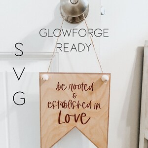 May include: A wooden banner sign with the text "be rooted & established in Love" in a dark brown script. The sign is suspended from a door knob with twine and small white decorative balls. The words "GLOWFORGE READY", "SVG", and "unstained designs" are also visible.