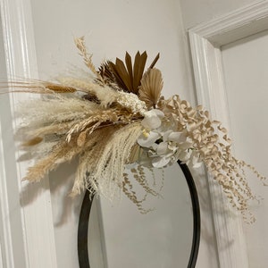 May include: A round mirror with a black frame is decorated with a dried floral arrangement. The arrangement features white and beige dried flowers, grasses, and palm leaves.