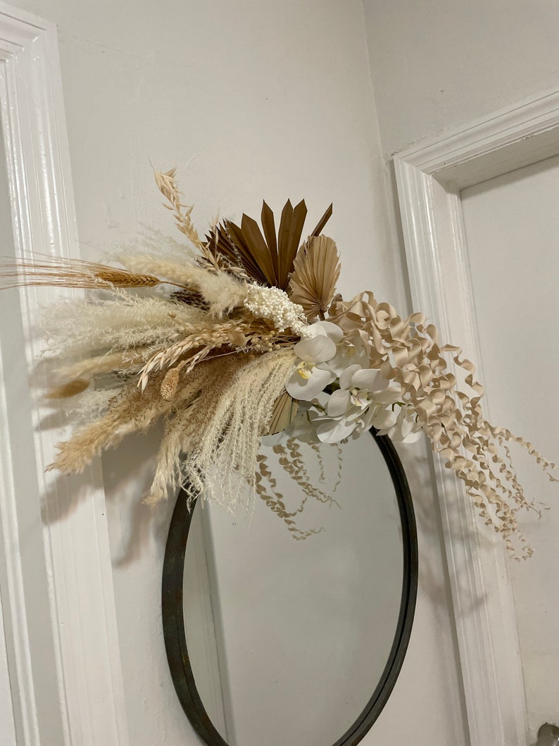 May include: A round mirror with a black frame is decorated with a dried floral arrangement. The arrangement features white and beige dried flowers, grasses, and palm leaves.