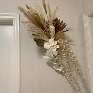 May include: A dried floral arrangement with white orchids, pampas grass, and dried palm leaves. The arrangement is a light beige and white color.