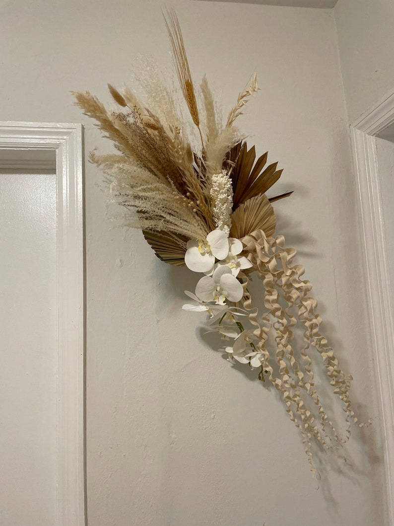 May include: A dried floral arrangement with white orchids, pampas grass, and dried palm leaves. The arrangement is a light beige and white color.