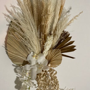 May include: A dried floral arrangement featuring white and beige pampas grass, dried palm leaves, and white orchids. The arrangement is perfect for adding a touch of natural beauty to any room.