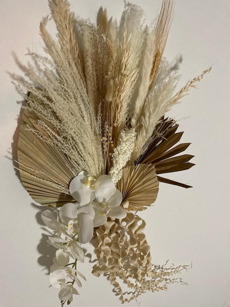 May include: A dried floral arrangement featuring white and beige pampas grass, dried palm leaves, and white orchids. The arrangement is perfect for adding a touch of natural beauty to any room.