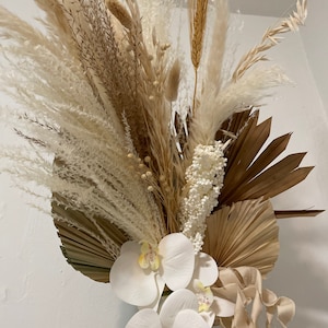 May include: A dried floral arrangement featuring white pampas grass, brown dried palm leaves, white orchids, and beige curly ribbon.