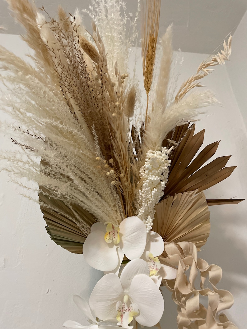 May include: A dried floral arrangement featuring white pampas grass, brown dried palm leaves, white orchids, and beige curly ribbon.