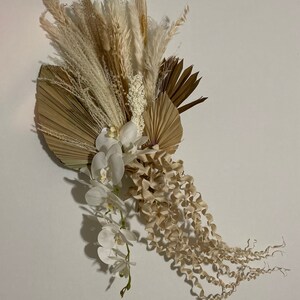 May include: A dried floral arrangement featuring white orchids, pampas grass, and dried palm leaves in shades of beige and brown.