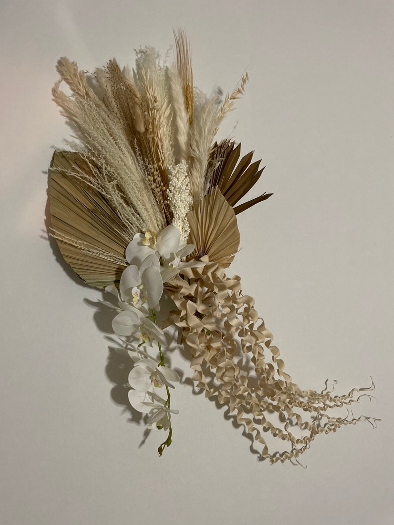 May include: A dried floral arrangement featuring white orchids, pampas grass, and dried palm leaves in shades of beige and brown.