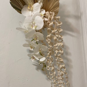 May include: A white orchid flower arrangement with beige curly tendrils hanging down. The orchids are arranged in a cascading style with the flowers facing outward. The background is a white wall.