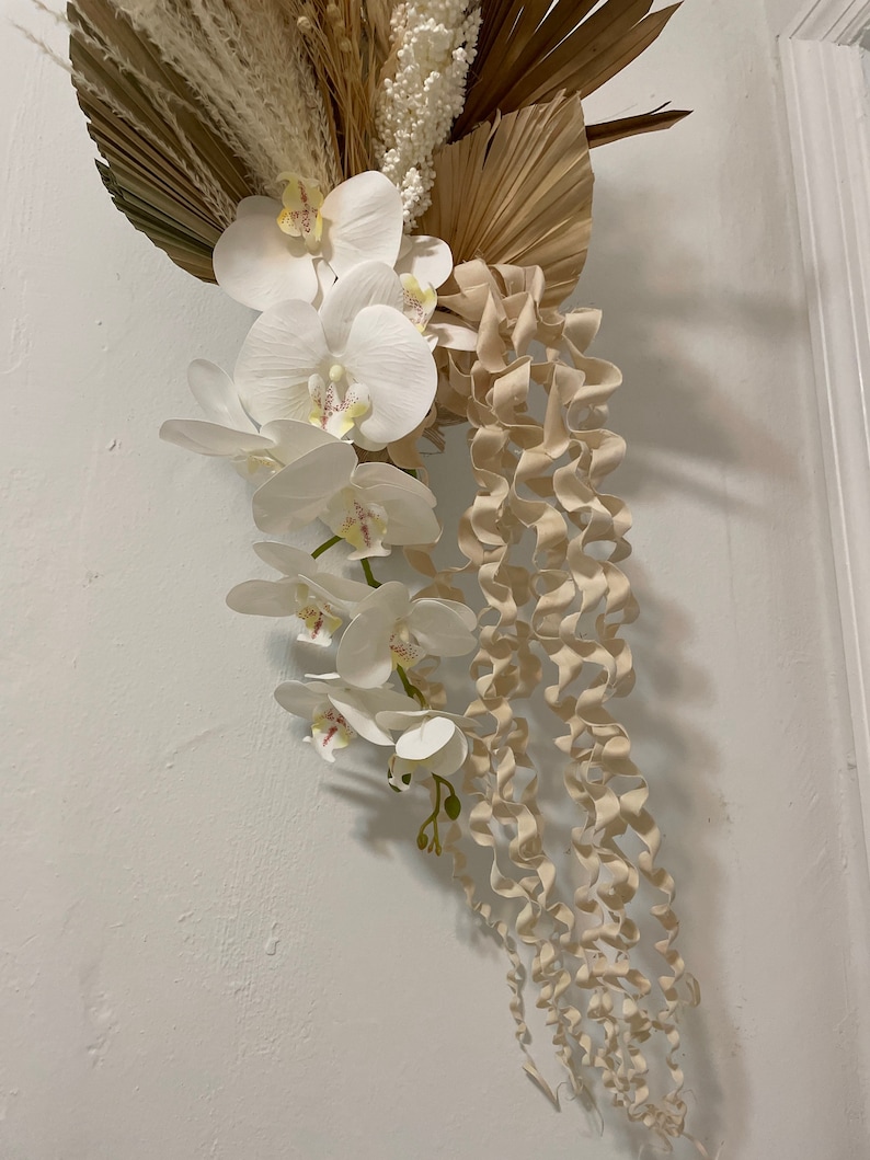 May include: A white orchid flower arrangement with beige curly tendrils hanging down. The orchids are arranged in a cascading style with the flowers facing outward. The background is a white wall.