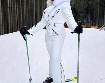 Puede incluir: Una persona con un traje de esquí blanco, casco y gafas de protección está de pie sobre esquís en la nieve, sosteniendo bastones de esquí. Los esquís son negros, amarillos y azules. El fondo muestra una ladera nevada y árboles.
