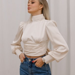 Close-up front view of the ivory bishop sleeve turtleneck blouse showing the gathered puff at the shoulder, full bishop sleeve with gathered cuff, fitted bodice, and smooth silk blend fabric; neutral background.