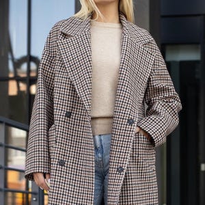May include: A double-breasted houndstooth coat in brown, black, and cream. The coat has a classic notched collar, two rows of buttons, and pockets. The model is wearing a beige sweater and blue jeans. The coat is a stylish outerwear option.