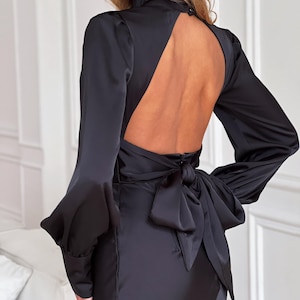 Może przedstawiać: Czarna satynowa sukienka z długimi rękawami i wysokim dekoltem. Sukienka ma dramatyczny odkryty tył i dużą kokardę w talii. Rękawy mają unikalny kształt dzwonu przy mankietach. Sukienka jest dopasowana i elegancka.