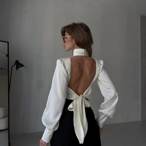 May include: A white satin blouse with long sleeves and a high neck, featuring an open back design tied with a bow. The blouse is paired with black trousers. The overall style is elegant and sophisticated.
