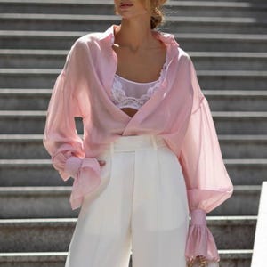 May include: A woman wearing a light pink, sheer button-down shirt with voluminous sleeves and white high-waisted pants. The shirt is paired with a white lace bralette. She is wearing square-framed glasses and carrying a small, cream-colored purse.
