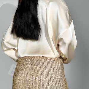 May include: A woman wearing a white satin blouse and a gold sequined skirt. The skirt is a midi length and has a fitted silhouette.