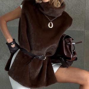 K&ouml;nnte beinhalten: Eine braune Kunstfellweste mit passendem Barett und Lederg&uuml;rtel. Das Outfit umfasst eine braune Lederhandtasche, schwarze Handschuhe und eine goldene Anh&auml;ngerkette. Das Model tr&auml;gt ein wei&szlig;es Oberteil und Shorts sowie schwarze Stiefel.