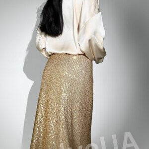 May include: A woman wearing a white satin blouse and a gold sequined skirt. The skirt is long and flowing, and the sequins are arranged in a vertical pattern. The woman is standing in front of a white background.