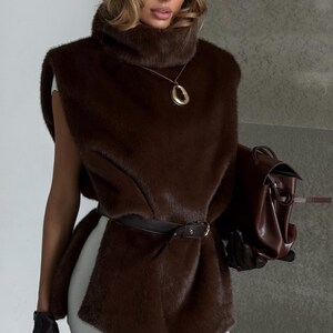 K&ouml;nnte beinhalten: Ein modisches Outfit mit einer braunen Kunstfellweste, einem passenden Barett und einer braunen Lederhandtasche. Das Outfit umfasst einen schwarzen G&uuml;rtel, Handschuhe und eine goldene Anh&auml;ngerkette. Der Look ist schick und stilvoll.