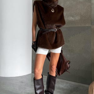 K&ouml;nnte beinhalten: Eine Frau tr&auml;gt eine braune Kunstfellweste, eine passende Baskenm&uuml;tze und kniehohe Stiefel. Das Outfit umfasst einen schwarzen G&uuml;rtel, wei&szlig;e Shorts und eine braune Lederhandtasche. Die Stiefel haben einen hohen Absatz.