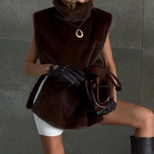 K&ouml;nnte beinhalten: Eine braune Kunstfellweste mit passendem Barett und hohen Stiefeln. Die Weste hat einen hohen Kragen und wird mit einer goldenen Anh&auml;ngerkette und einer braunen Lederhandtasche kombiniert. Das Barett hat ein goldenes Symbol.