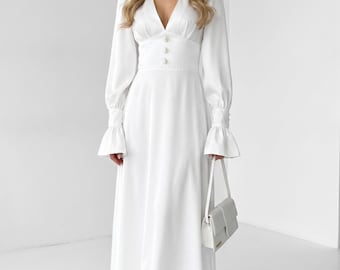 May include: A white dress with a V-neckline, long sleeves, and a flared skirt. The dress features button detailing at the waist and is paired with a white handbag and nude heels. The dress is a formal style.
