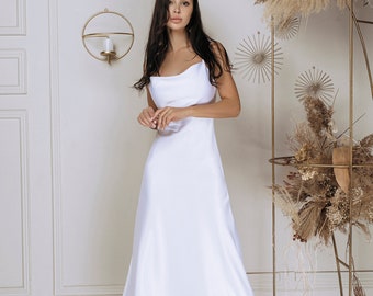 May include: A white satin midi dress with a cowl neckline. The dress is paired with black heels with silver pointed toes. The background features a decorative gold metal structure with dried floral arrangements.