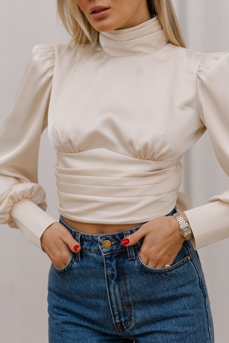 Three-quarter front view of the ivory silk blend turtleneck blouse showing the high neck, bishop sleeves with slight shoulder puff, and fitted cropped bodice; neutral background.